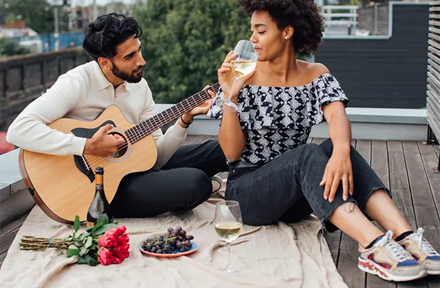 A couple enjoying a picnic on a blanket, strumming a guitar and savoring a glass of wine.