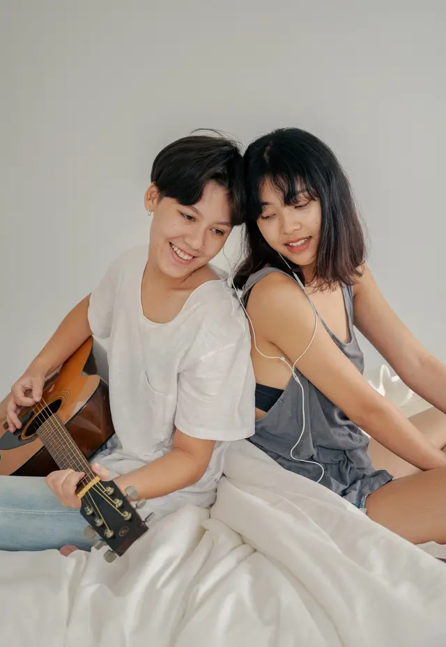 Two young Asian women sitting on a bed, strumming a guitar, radiating love and harmony.