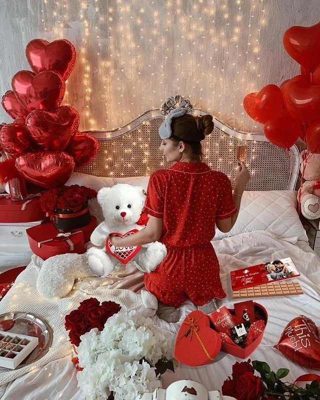 A woman sitting on a bed surrounded by Valentine's Day decorations, holding a teddy bear.