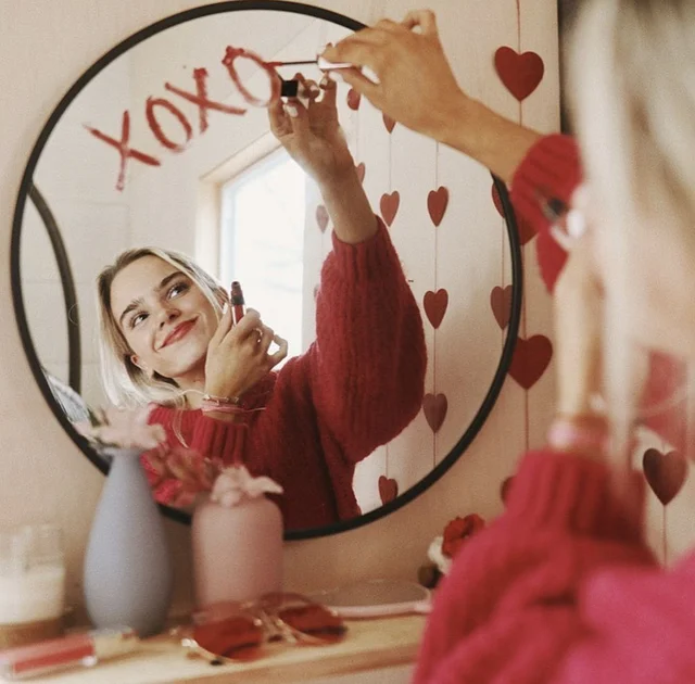 A woman in a red sweater applying lipstick in front of a mirror.