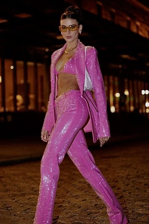 A stylish woman in a pink suit struts confidently on a cobblestone street, exuding power and sass with her bold fashion choices.
