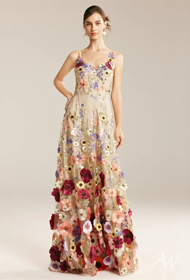 A young woman wearing a long, flowing floral dress stands tall. The dress features a vibrant pattern of large red, pink, white, and purple blooms scattered across a beige background fabric.