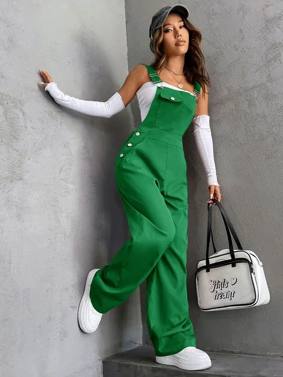 A woman in bright green jumpsuit with white buttons, wearing white long sleeves underneath, holding a white bag with black outlines, leaning against a textured grey wall.