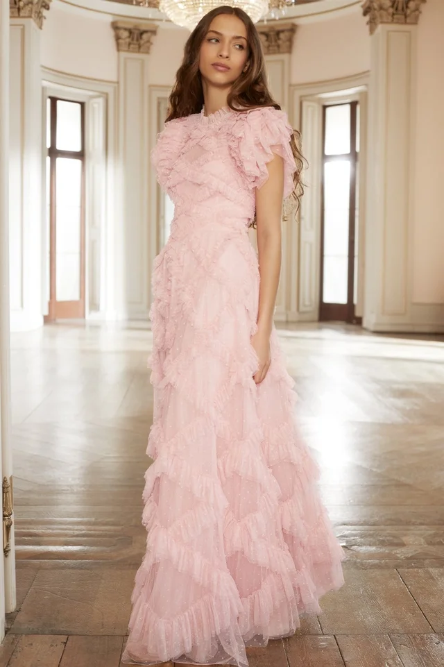A woman modeling a light pink frilly tulle prom dress with intricate floral appliques in an elegant ballroom setting.