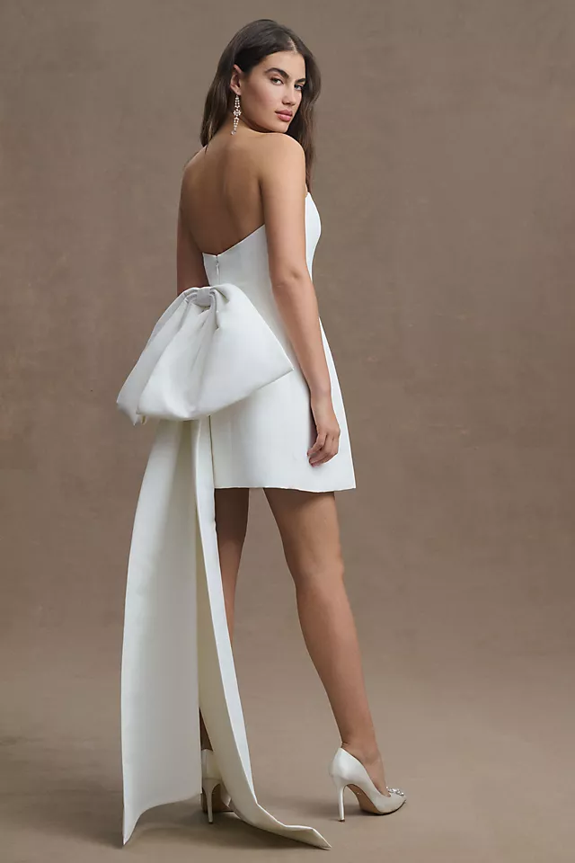 A chic strapless white mini dress with a pleated waistline and dramatic back bow detail, perfect for the modern bride