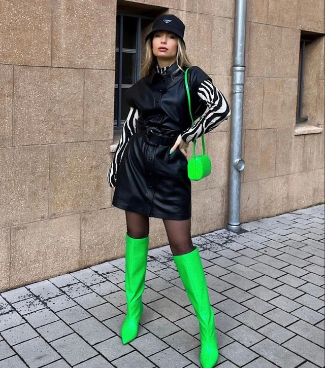 A woman in black leather dress with white stripes on sleeves, wearing bright green knee-high boots and holding a matching small round bag, standing on a sidewalk next to a brown stone building.