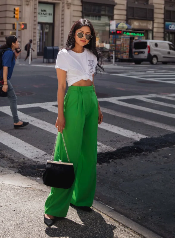 A woman in white cropped t-shirt and bright green pants, holding a small black purse, standing on a zebra crossing in an urban setting with buildings and traffic in the background.