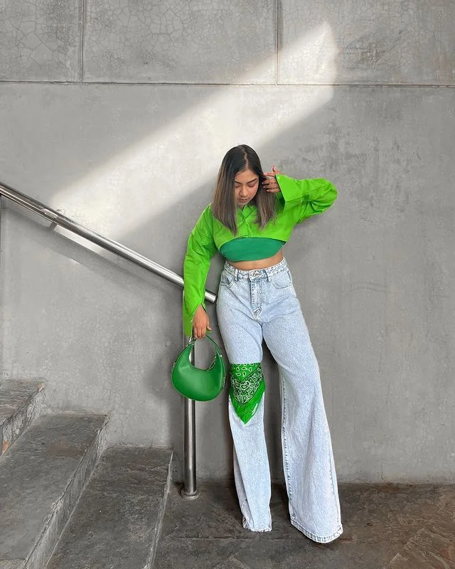 A stylish woman stands against a grey concrete wall, wearing a vibrant green cropped sweatshirt and light blue wide-leg jeans.