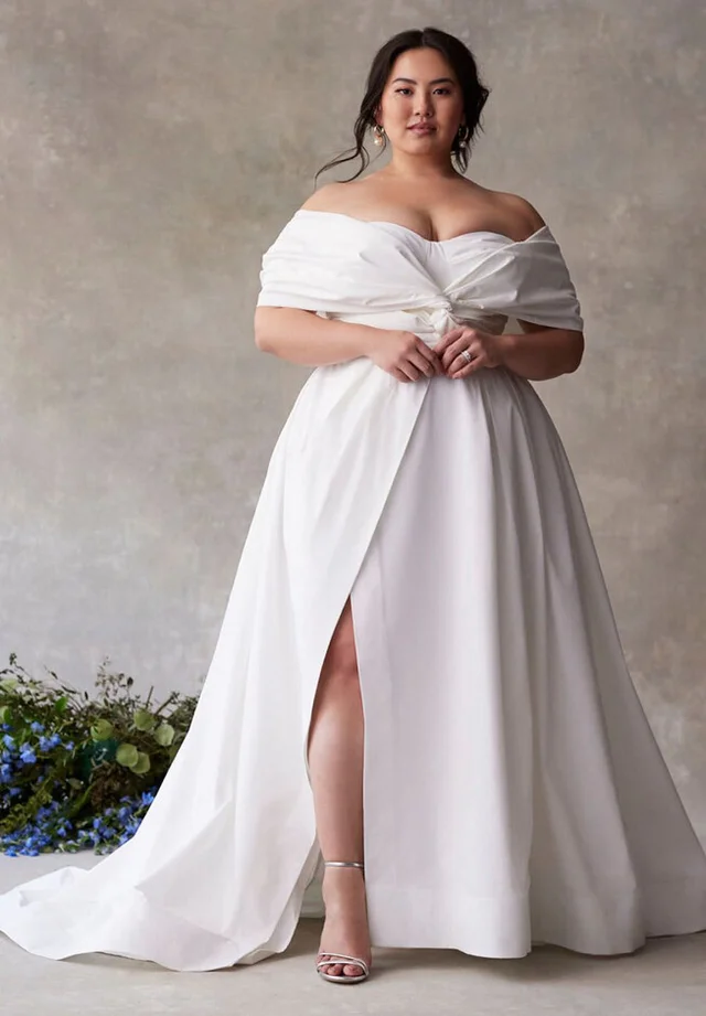 A curvy model poses confidently in a white off-the-shoulder satin wedding gown with a dramatic thigh-high slit