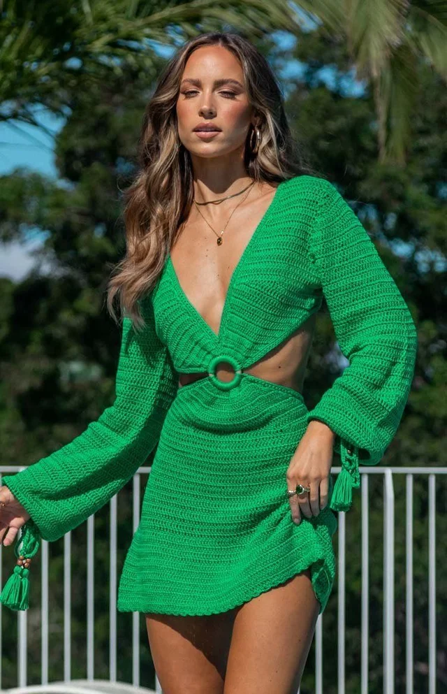 Young woman posing outdoors in a bright green crocheted dress with long sleeves and a deep neckline, accessorized with necklaces and rings.