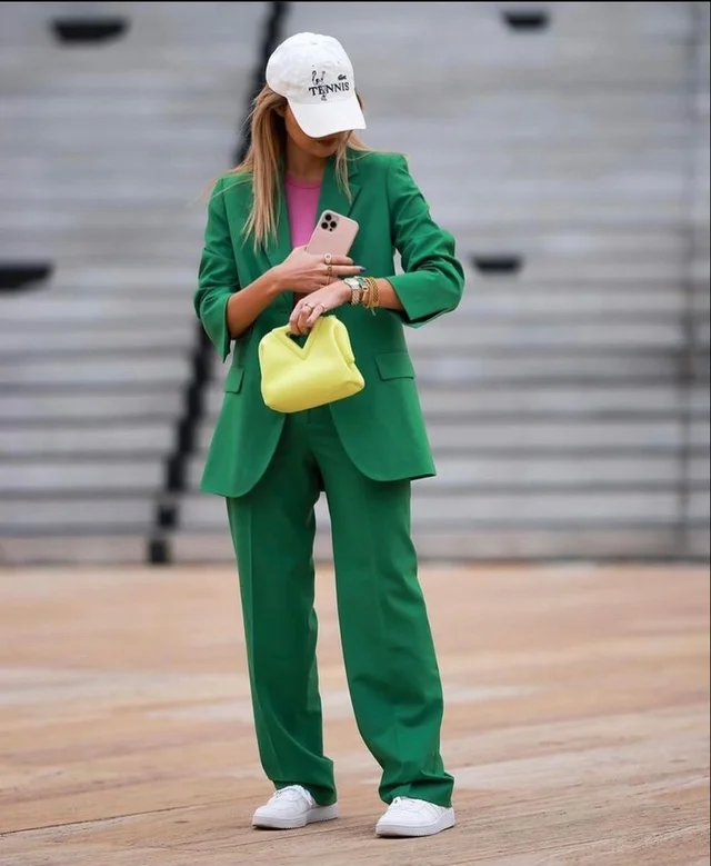 A woman wearing an emerald green pantsuit and white baseball cap walks while holding a yellow purse and looking at her phone.
