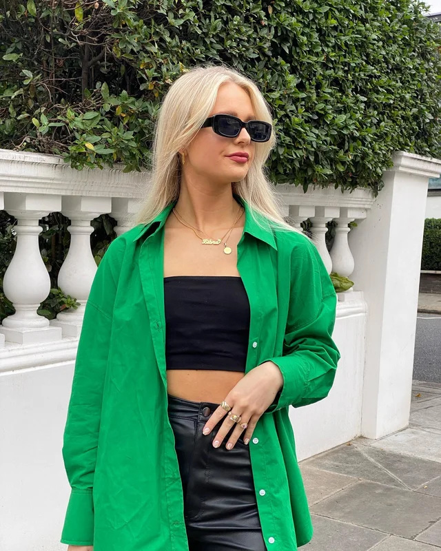 A blonde woman wearing a bright green button-up shirt over a black crop top and leather skirt poses in front of a hedge wall and decorative columns. She has sunglasses and a necklace on, and her hair is styled straight.