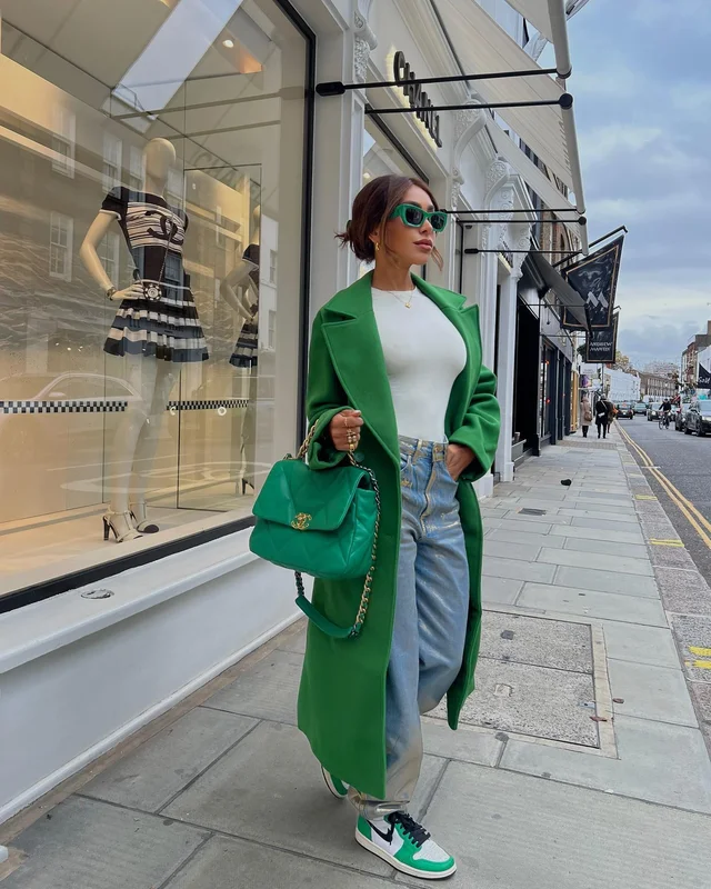 Stylish woman in bright green coat and matching bag standing on a sidewalk in front of a fashion store with mannequins displayed in the window.