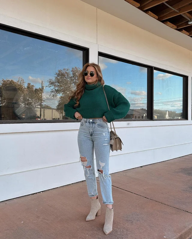 A woman wearing a green turtleneck sweater, distressed blue jeans, and beige boots stands on a patio or balcony area with large windows overlooking an outdoor scene.