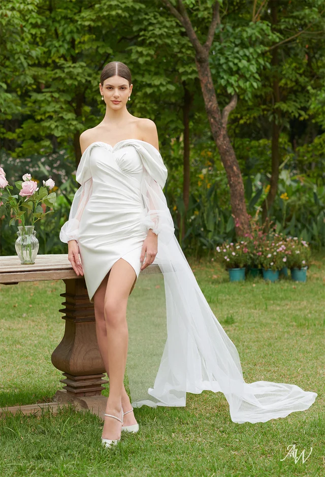 a young woman wearing an elegant off-the-shoulder white gown in a beautiful garden setting with trees and flowers