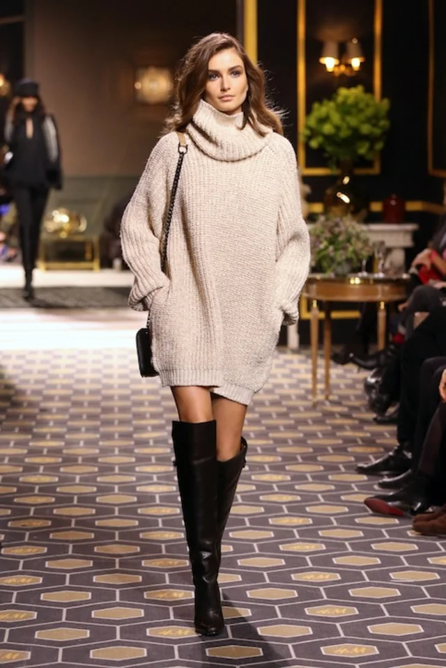 a woman in a turtleneck sweater dress and boots walking down a runway