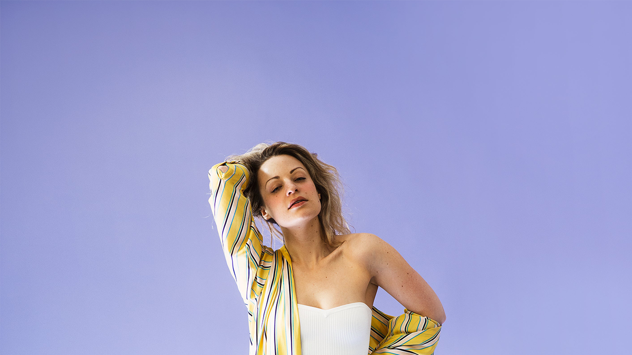 Woman stands with her hand in her hair, the striped yellow folds of her silky long summer fashion shirt flowing down. A great fashion pairing with her silky sheer loose summer pants.